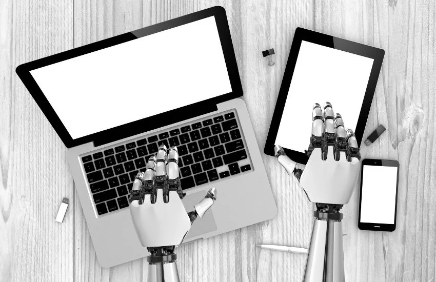 Manos robóticas utilizando un ordenador portátil, una tablet y un smartphone sobre mesa de madera, concepto de sistemas de IA prohibidos y regulación.